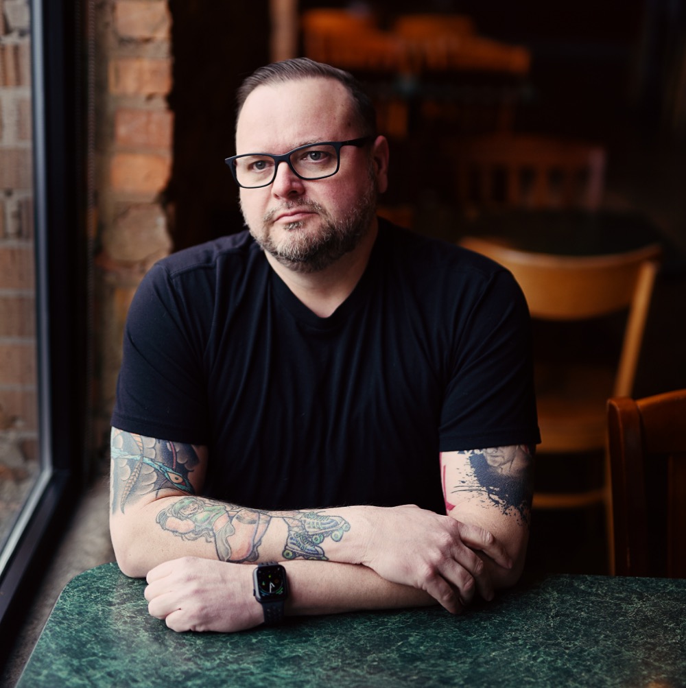 RyanKnott Ryan Knott, founder and owner of Secret Chord Marketing, sits at a green marble table in a modern café or restaurant setting. He's wearing a black v-neck t-shirt and black-rimmed glasses, with his arms crossed and resting on the table. Ryan has a well-groomed beard and colorful tattoo sleeves on both arms featuring floral and artistic designs. He's wearing a black smartwatch and has a warm, approachable expression. The background shows exposed brick walls and wooden chairs, creating a casual yet professional atmosphere that reflects his authentic, down-to-earth approach to content marketing consulting.