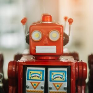 A vintage-style red toy robot with a rectangular head featuring two large circular yellow eyes and a white rectangular mouth. The robot has small antennae protruding from the top of its head and displays two blue analog gauges with yellow dials on its chest. The image shows a close-up view of the robot's upper body against a soft, blurred background.