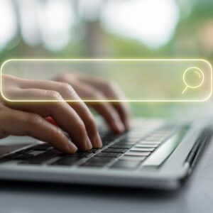 Hands typing on a laptop keyboard with a translucent search bar overlay featuring a magnifying glass icon, representing online search and SEO concepts. The image has a soft, blurred green background suggesting an outdoor or natural setting, symbolizing the digital search process and search engine optimization strategies used in content marketing.