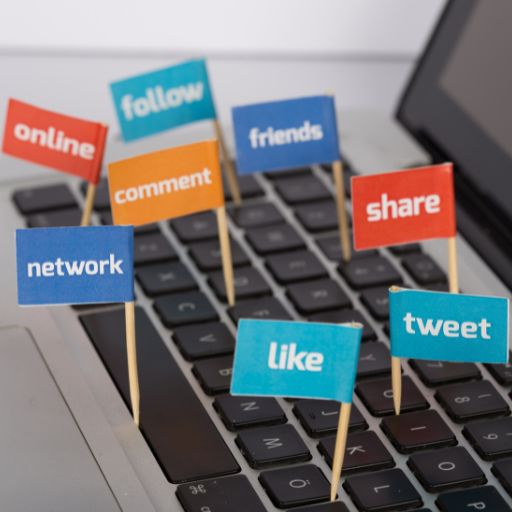 A laptop keyboard with small colorful paper flags on toothpicks displaying social media and digital marketing terms including "online," "follow," "friends," "comment," "share," "network," "like," and "tweet." The flags are in various bright colors - red, blue, teal, and orange - representing the diverse landscape of social media platforms and digital engagement strategies used in content marketing and social media consulting.